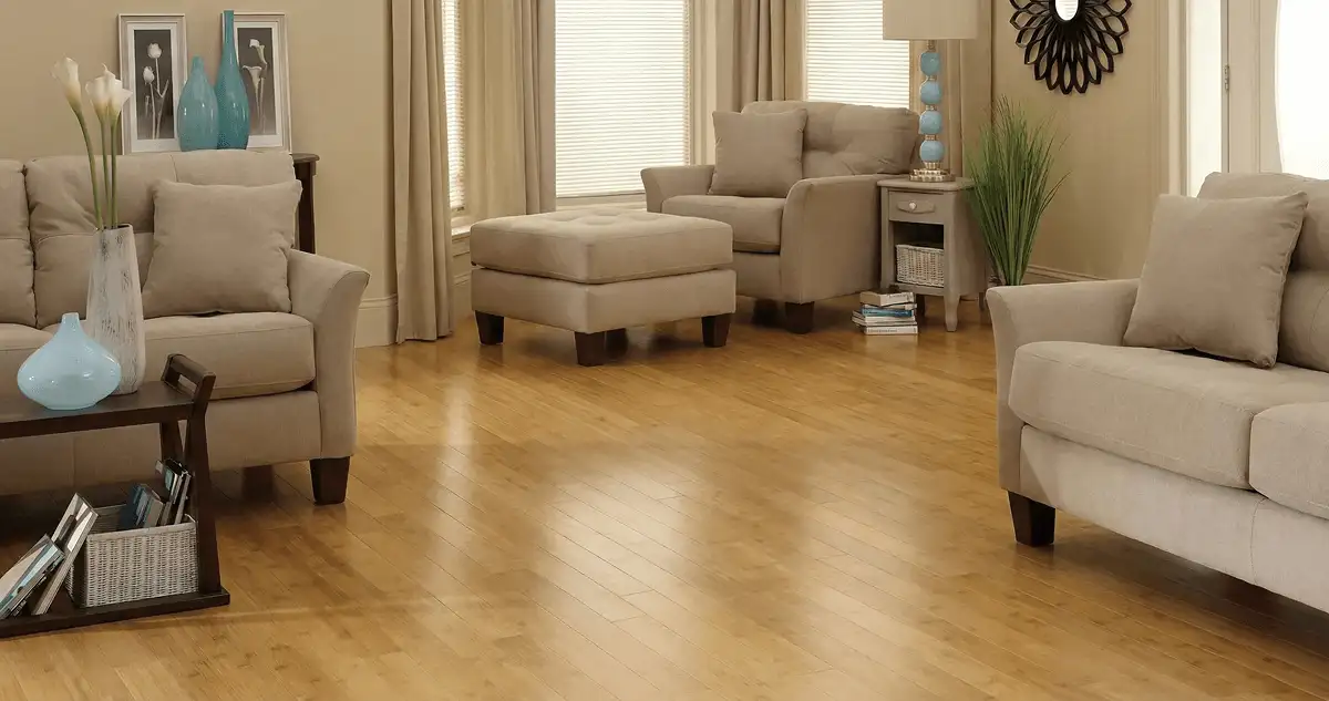 A living room with professionally installed bamboo flooring, beige sofas, a matching ottoman, a side table with a blue lamp, vases, framed pictures, and large windows with blinds and curtains in Charlotte, NC